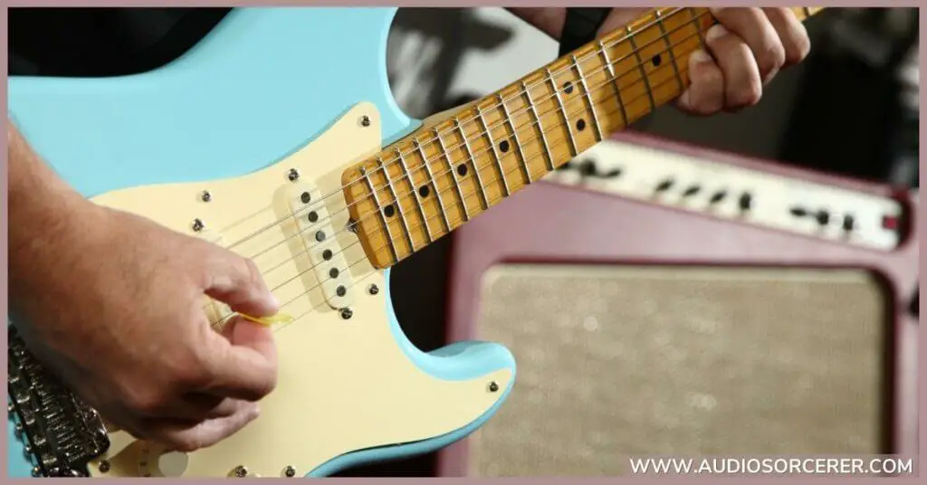 Closeup of a guy playing a blue Fender Stratocaster with an amp in the background.