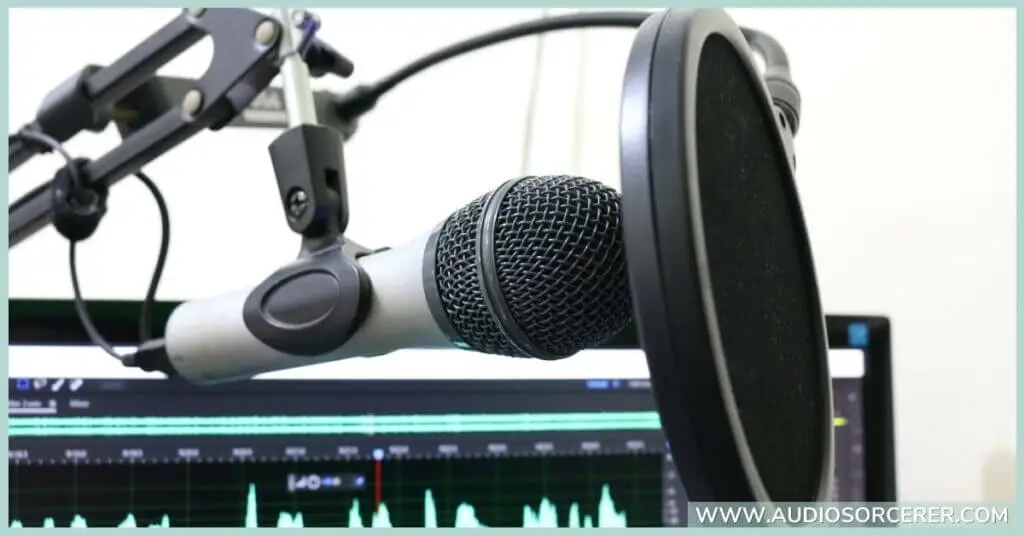 Microphone in a podcast studio with a pop filter in front of it and a computer in the background showing an audio waveform.